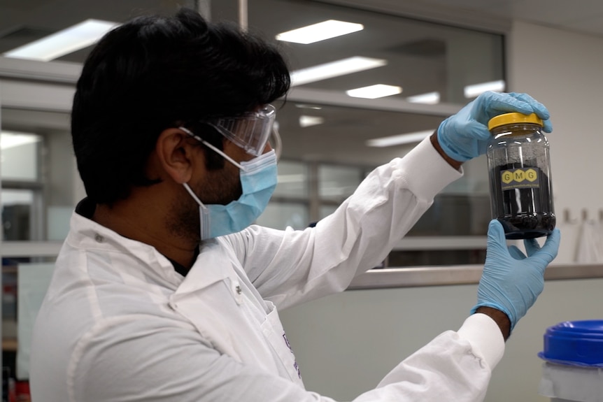 Man in a white lab coat holding a jar with yellow lid and dark-coloured chemicals in it.