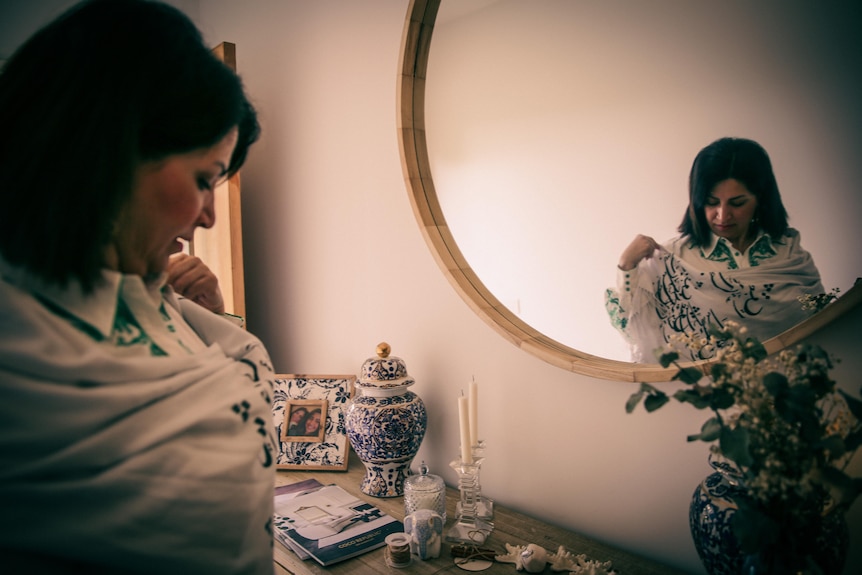 A woman stands in front a mirror wearing a white sheath over clothes which is printed with Arabic writing