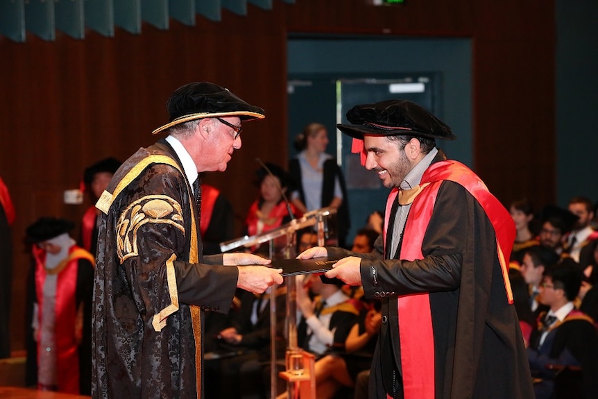 A man dressed in a University gown and hat receives his PhD from another a man in a University gown and hat.