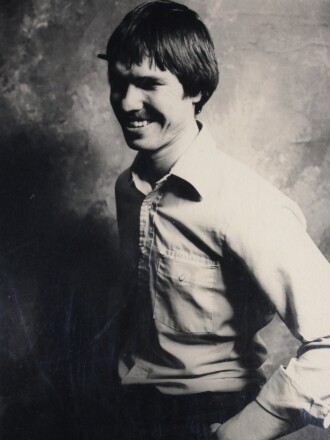 Photo of a man with longer hair, a  moustache, and a pencil behind his ear,  age 22, standing in a black-and-white photo against a portrait backdrop.