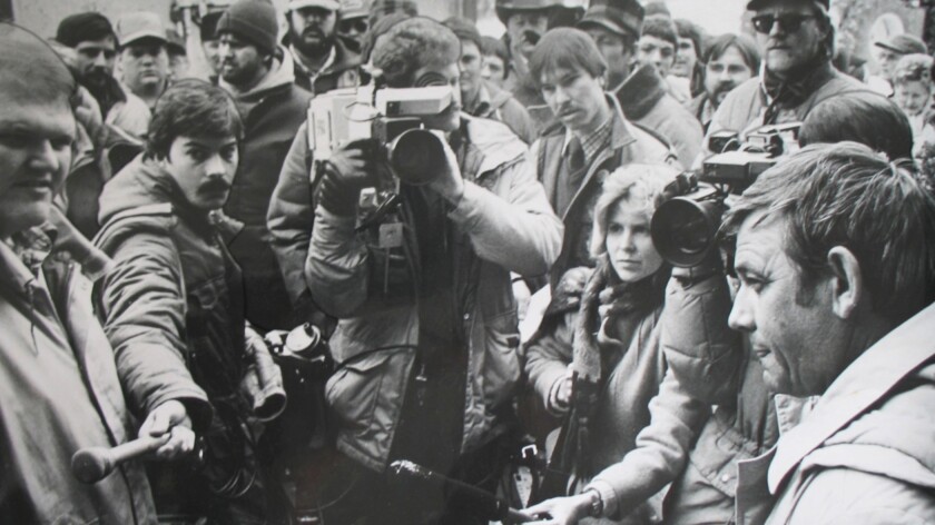 A scrum of news reporters and farmers at a farm protest, while a bank board member listens at right.