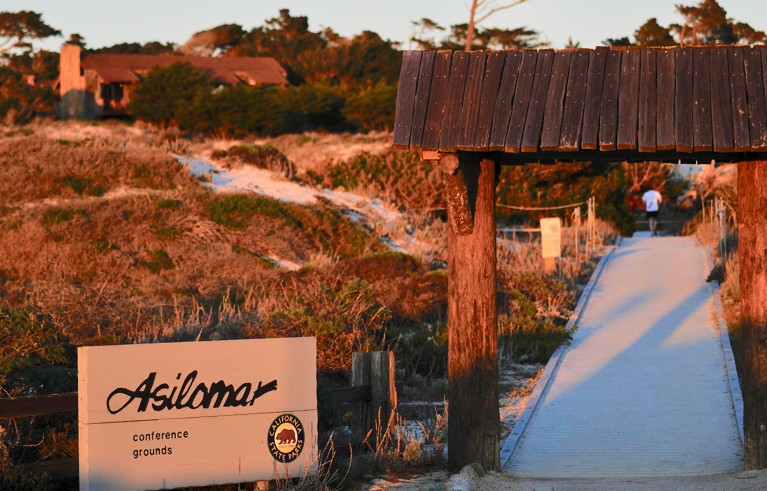 Sunset shot of a the outdoor space of a conference site. Garden, paved walkpath and wood entrance is on the left and sign saying Asilomar is on the left.